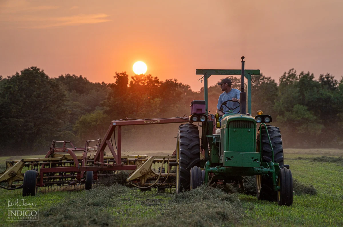 Farmer raking hay with a tractor at sunset.